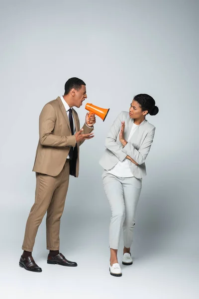 Hombre de negocios afroamericano enojado gritándole a la mujer de negocios mientras sostiene el megáfono en gris - foto de stock
