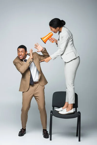 Angry african american businesswoman standing on chair and screaming at scared businessman while holding megaphone on grey — Stock Photo