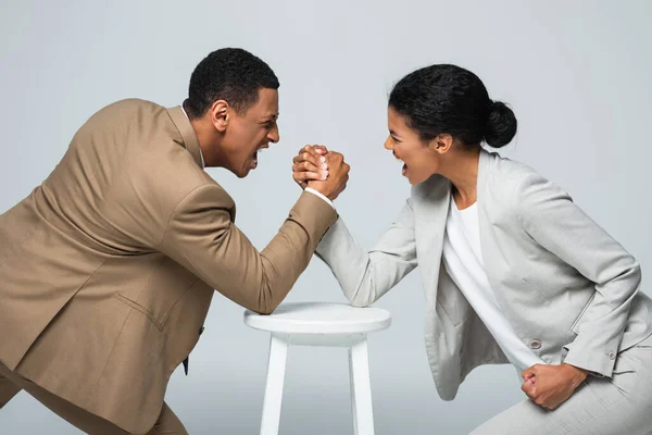 Vista lateral de hombre y mujer de negocios afroamericana haciendo lucha libre de brazos en silla blanca aislada en gris - foto de stock