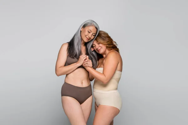 Mujeres sonrientes multiétnicas en ropa interior cogidas de la mano aisladas en gris, concepto positivo para el cuerpo - foto de stock