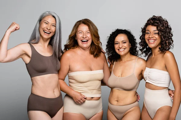 Asian woman in underwear showing muscles near cheerful interracial friends isolated on grey — Stock Photo