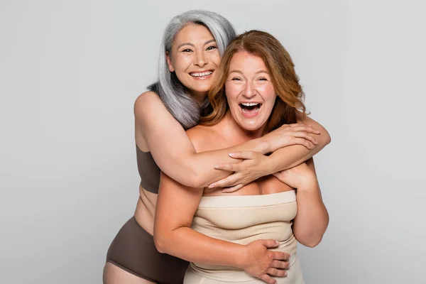 Sonriente asiático mujer en ropa interior abrazo amigo aislado en gris - foto de stock