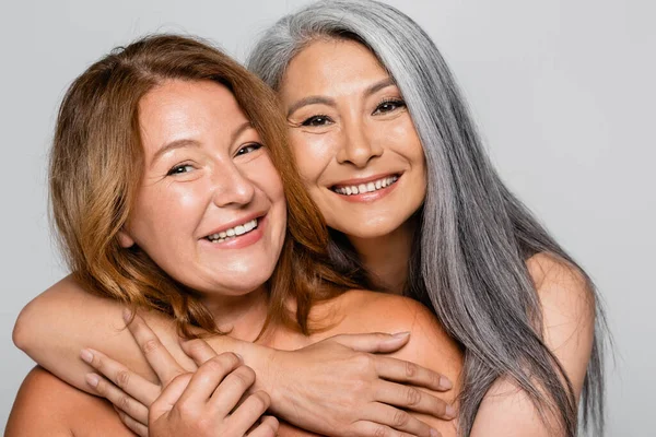 Smiling asian woman hugging friend with naked shoulders isolated on grey — Stock Photo