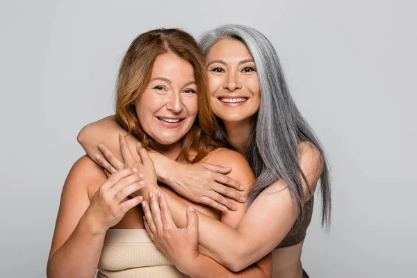Sorridente donna asiatica abbracciare amico in reggiseno isolato su grigio, corpo concetto positivo — Foto stock