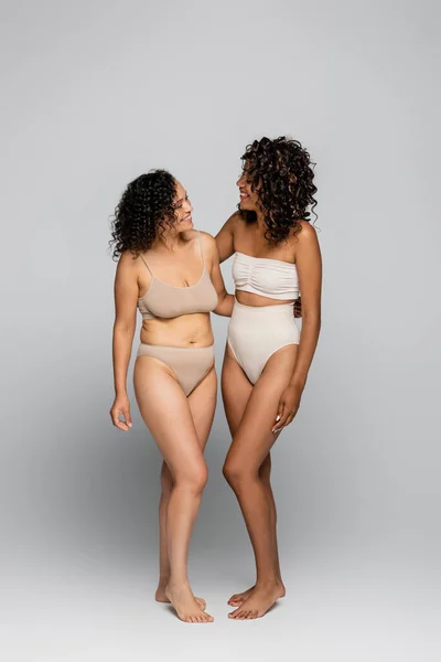 Sonrientes mujeres afroamericanas en lencería abrazándose sobre fondo gris, concepto positivo para el cuerpo - foto de stock