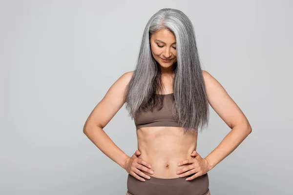 Mujer asiática en ropa interior cogida de la mano en las caderas aislada en gris, concepto positivo para el cuerpo - foto de stock