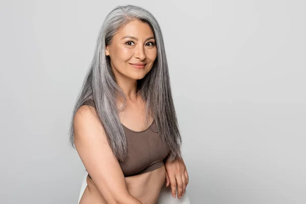 Asiática mujer en sujetador sentado en silla y sonriendo a la cámara aislado en gris - foto de stock