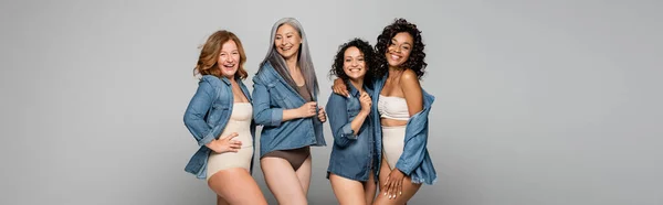 Позитивные многоэтнические женщины в нижнем белье и джинсовой рубашке изолированы на сером, баннер — стоковое фото