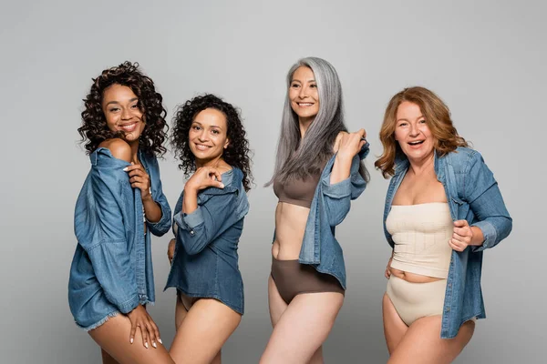 Mujeres sonrientes multiétnicas en ropa interior ajustando camisas de mezclilla aisladas en gris - foto de stock