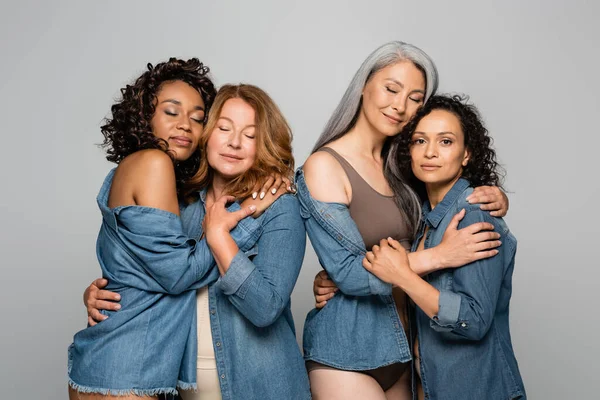 Mujeres multiculturales en camisas vaqueras abrazándose aisladas en gris - foto de stock