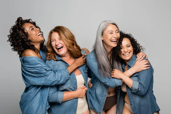 Glückliche multiethnische Frauen in Unterwäsche und Jeanshemden, die sich isoliert auf grauem, körperpositivem Konzept umarmen — Stockfoto