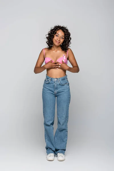 Smiling african american woman in bra with ribbon of breast cancer awareness on grey background — Stock Photo