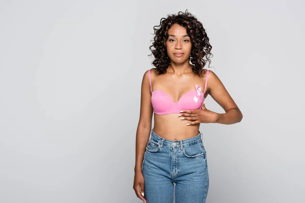 African american woman touching pink bra with ribbon of breast cancer awareness isolated on grey — Stock Photo