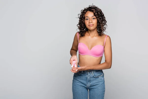 Молода афроамериканська жінка з бюстгальтером, що тримає в руках стрічку інформованості про рак молочної залози ізольовану на сірому — стокове фото
