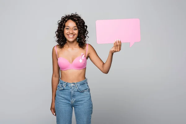 Mujer afroamericana sonriente con cinta de conciencia de cáncer de mama sosteniendo burbuja del habla aislada en gris - foto de stock