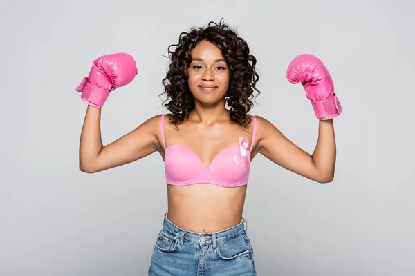 African american woman in boxing gloves and bra with pink ribbon smiling isolated on grey — Stock Photo