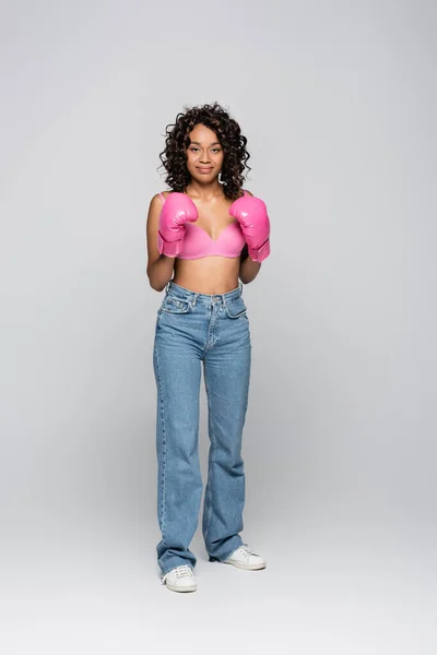 Sorridente donna afroamericana in reggiseno rosa e guanti da boxe su sfondo grigio — Foto stock