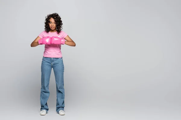 Jeune femme afro-américaine en t-shirt avec ruban et gants de boxe sur fond gris — Photo de stock