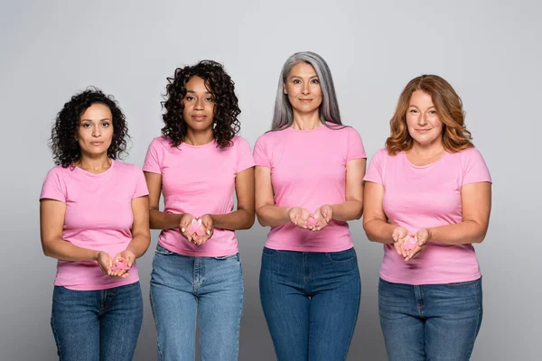 Багатонаціональні жінки, що тримають стрічки усвідомлення раку молочної залози, дивляться на камеру на сірому фоні — стокове фото