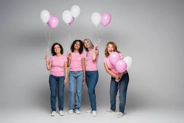 Mujeres sonrientes multiétnicas con cintas rosas y globos sobre fondo gris - foto de stock