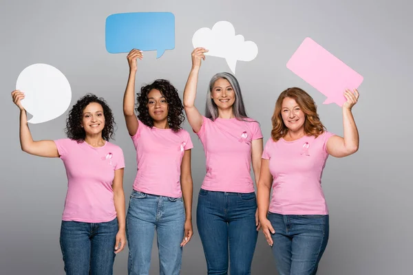 Lächelnde multiethnische Frauen mit rosa Schleifen, die Sprechblasen auf grauem Hintergrund halten — Stockfoto