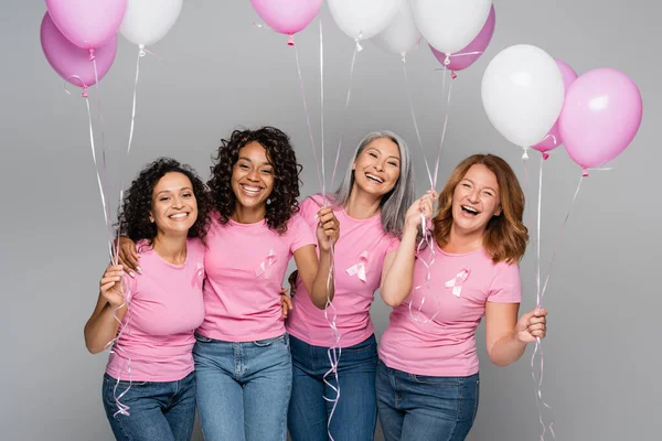 Mujeres alegres multiétnicas con cintas de conciencia de cáncer de mama sosteniendo globos aislados en gris - foto de stock