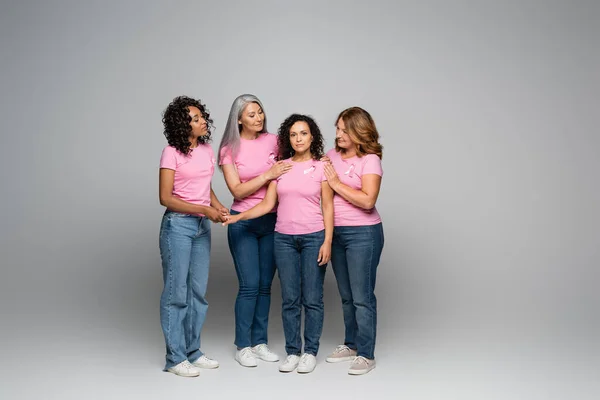 Multiethnic women hugging friend with pink ribbon on t-shirt on grey background — Stock Photo