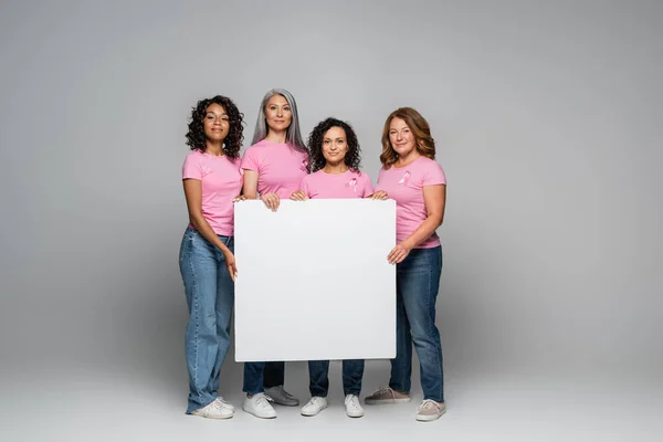 Mulheres multiétnicas com fitas de consciência de câncer de mama segurando cartaz em fundo cinza — Fotografia de Stock