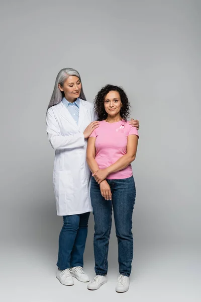 Souriant asiatique médecin câlin afro-américaine femme avec ruban de sensibilisation au cancer du sein sur fond gris — Photo de stock