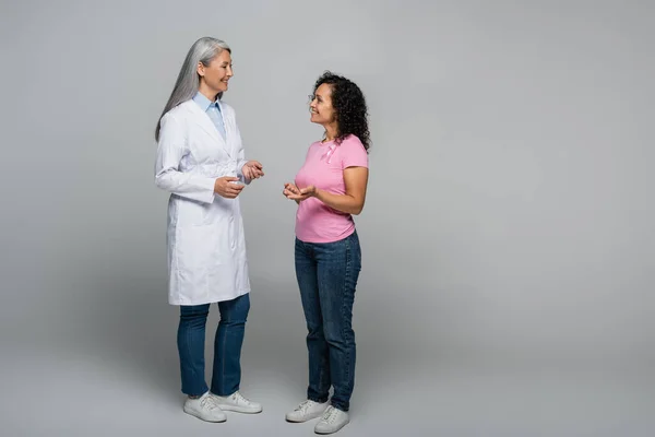 Веселий азійський лікар, який розмовляє з пацієнтом - афроамериканцем зі стрічкою усвідомлення раку молочної залози на сірому тлі. — стокове фото