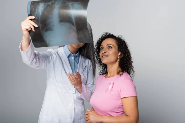 Doctor apuntando a la fluorografía cerca de la sonriente mujer afroamericana con cinta rosa aislada en gris - foto de stock