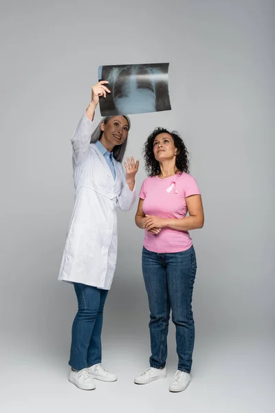 Sourire asiatique médecin pointant fluorographie près afro-américaine femme avec ruban de la sensibilisation au cancer du sein isolé sur gris — Photo de stock