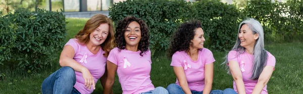 Alegres mujeres multiculturales con cintas rosadas sentadas en la hierba, pancarta - foto de stock