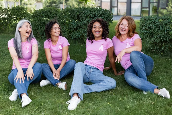 Mujeres sonrientes multiétnicas en camisetas rosas con cintas sentadas en la hierba - foto de stock