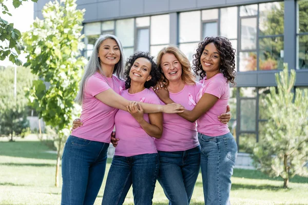 Smiling multiethnic women with ribbons of breast cancer awareness hugging outdoors — Stock Photo