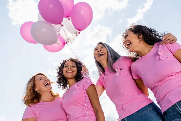 Niedriger Blickwinkel auf glückliche multiethnische Frauen mit Brustkrebsbändern und Luftballons im Freien — Stockfoto