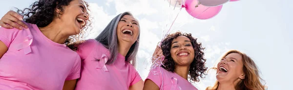 Low angle view of cheerful multiethnic women with pink ribbons and balloons outdoors, banner — Stock Photo