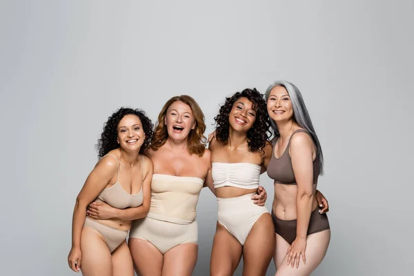 Cuerpo multiétnico mujeres positivas en lencería abrazando y mirando a la cámara aislada en gris - foto de stock