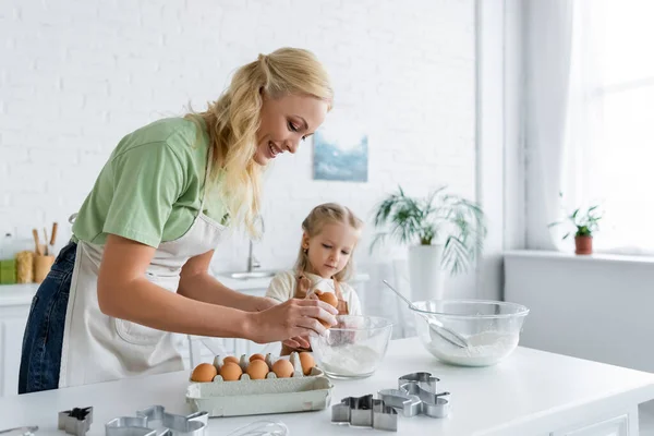 Mulher quebrar ovo em tigela enquanto cozinha com a filha na cozinha — Fotografia de Stock