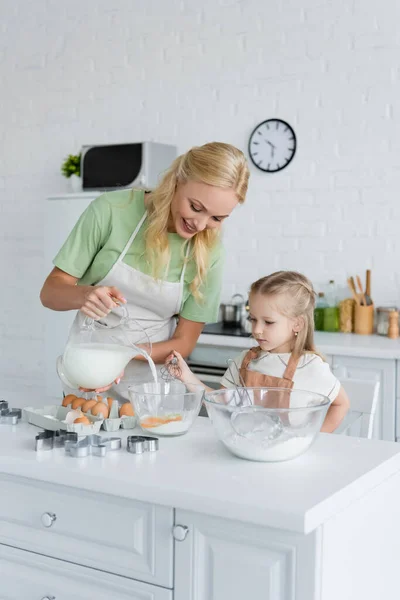 Smiling woman pouring milk into bowl near daughter with whisk — Stock Photo