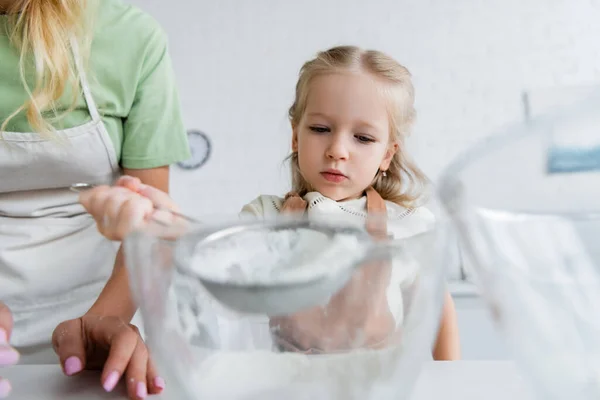 Kind sieben Mehl in Schüssel neben Mutter in Küche, verschwommener Vordergrund — Stockfoto