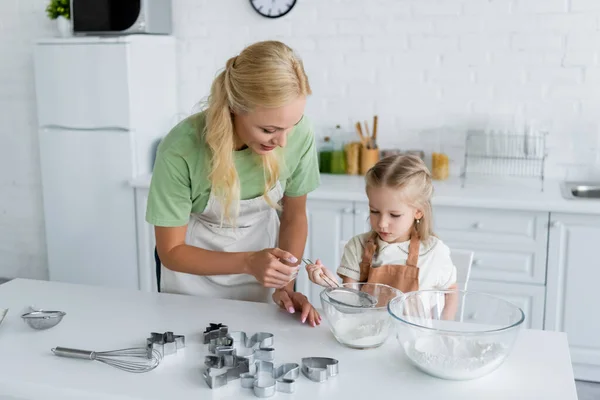 Chica tamizar harina en un tazón cerca de mamá y cortadores de galletas en la mesa de la cocina - foto de stock