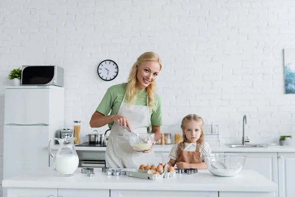 Cheerful woman mixing ingredients with whisk near little daughter in kitchen — Stock Photo