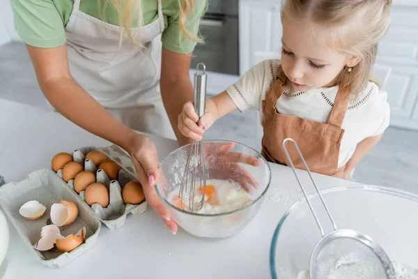 Kind mischt Hühnereier und Mehl mit Schneebesen in der Nähe der Mutter — Stockfoto