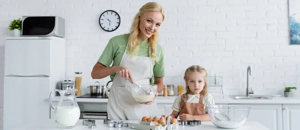 Sonriente madre mezclando ingredientes en un tazón mientras cocina con su hija en la cocina, pancarta - foto de stock