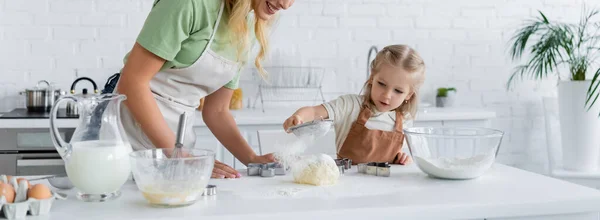 Niña tamizar la harina en la masa cerca de la madre sonriente y los ingredientes en la mesa, pancarta - foto de stock