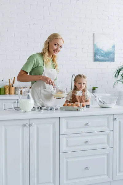 Счастливая мама смотрит в камеру, смешивая ингредиенты в миске рядом с дочерью — стоковое фото