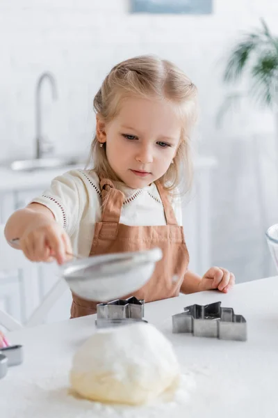 Ребенок в фартуке просеивает муку возле кусачки для печенья, размытый передний план — стоковое фото