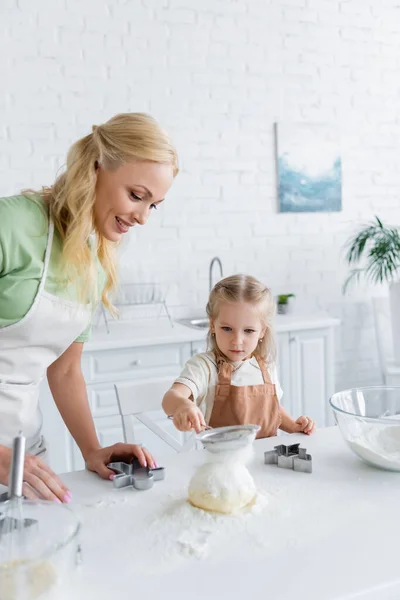 Criança peneirando farinha na massa crua enquanto ajuda a mãe na cozinha — Fotografia de Stock