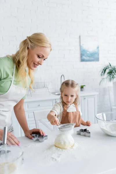 Niño tamizar la harina en masa cruda mientras ayuda a mamá en la cocina - foto de stock